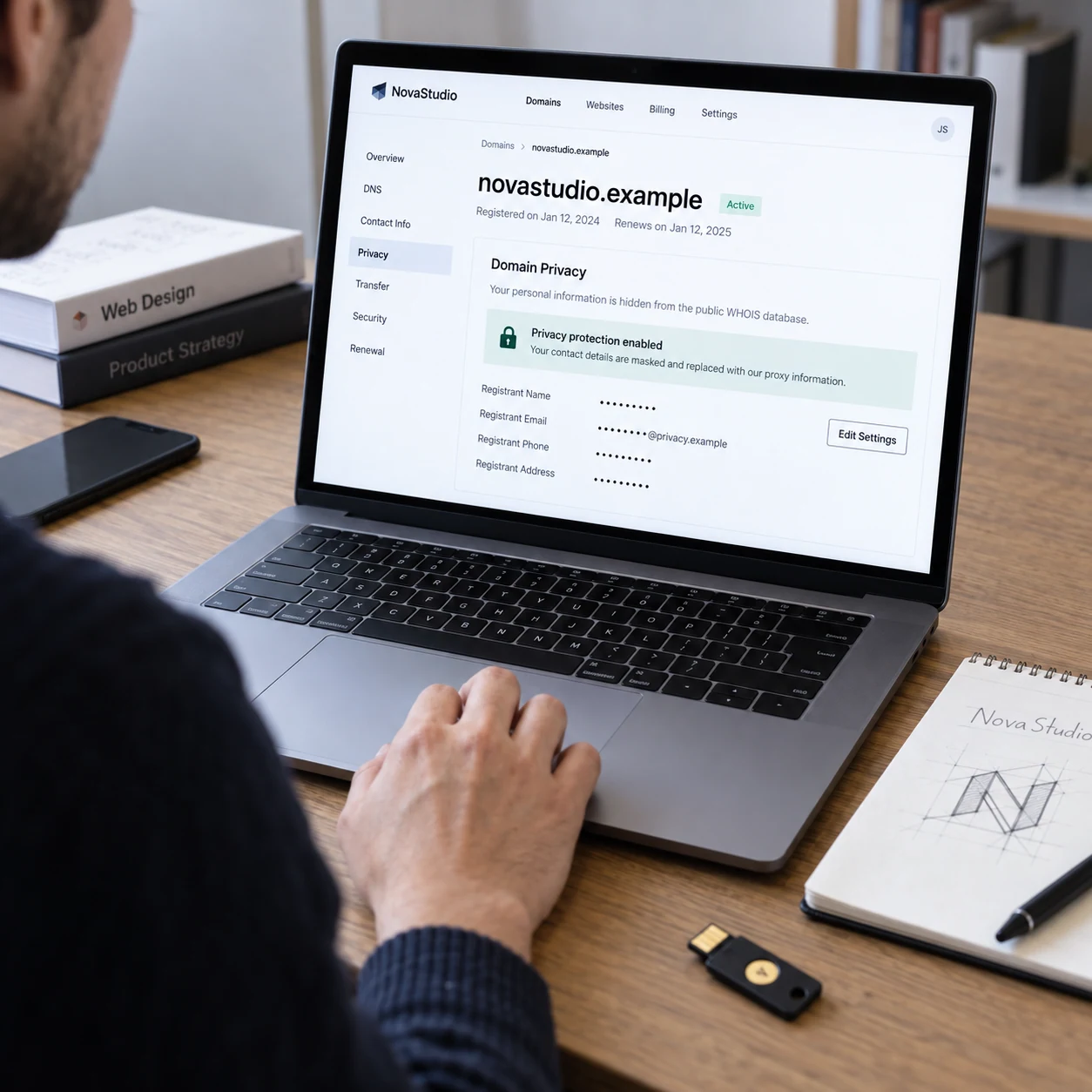 Close photo of laptop displaying a domain privacy overview for fictional 'novastudio.example', personal contact fields visibly masked, physical notebook with brand sketch and a security key nearby, hand on trackpad