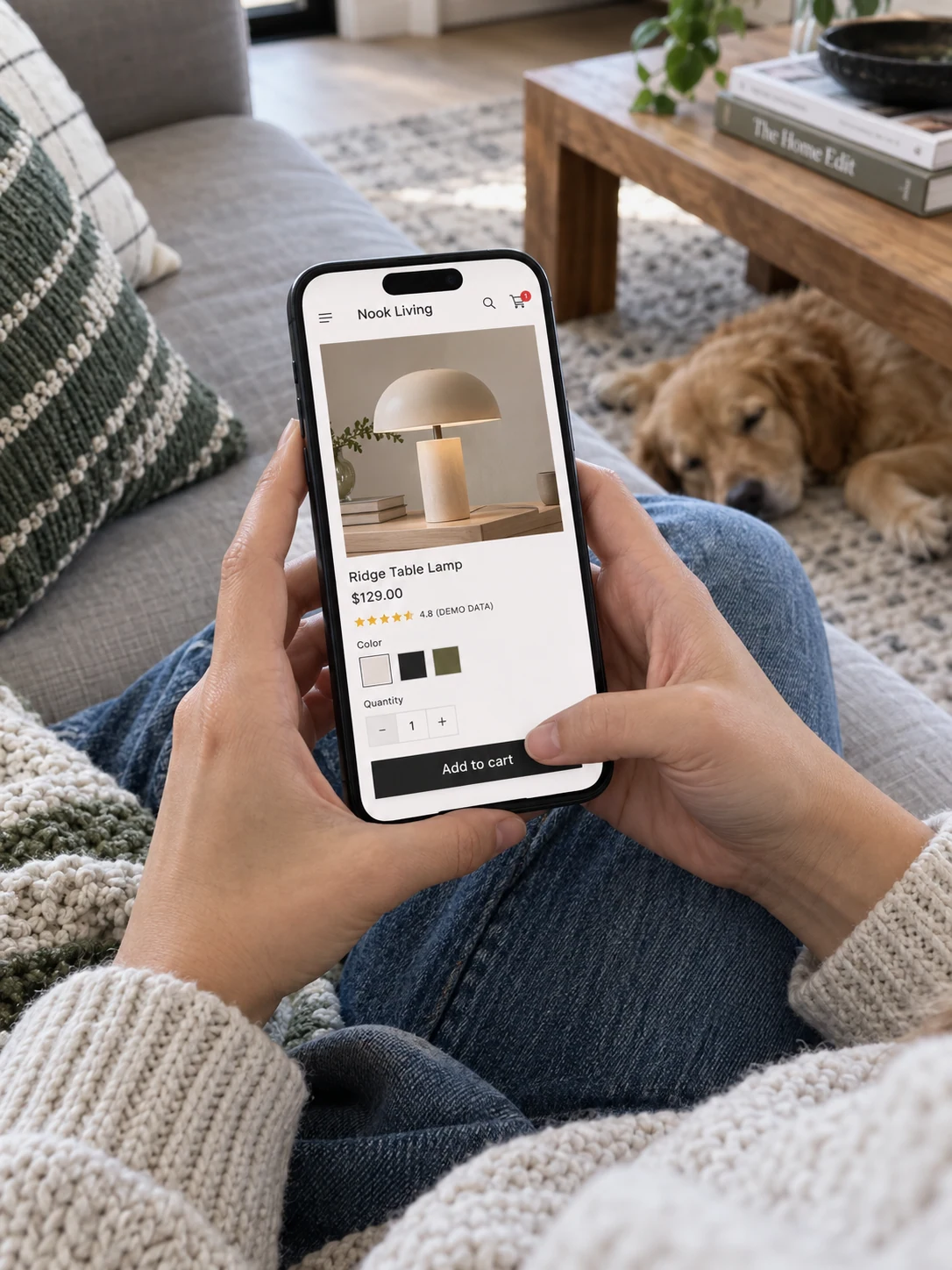 A shopper seated on sofa uses a smartphone supported naturally by both palms, screen facing viewer shows fictional 'Nook Living' lamp added to shopping cart