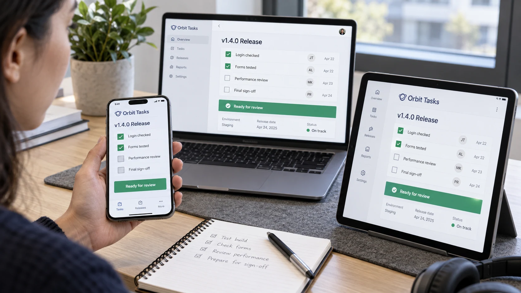 Quality tester at desk with phone in a LEFT hand and tablet on stand, both display fictional 'Orbit Tasks' release checklist, clear 'Login checked', 'Forms tested', 'Ready for review'