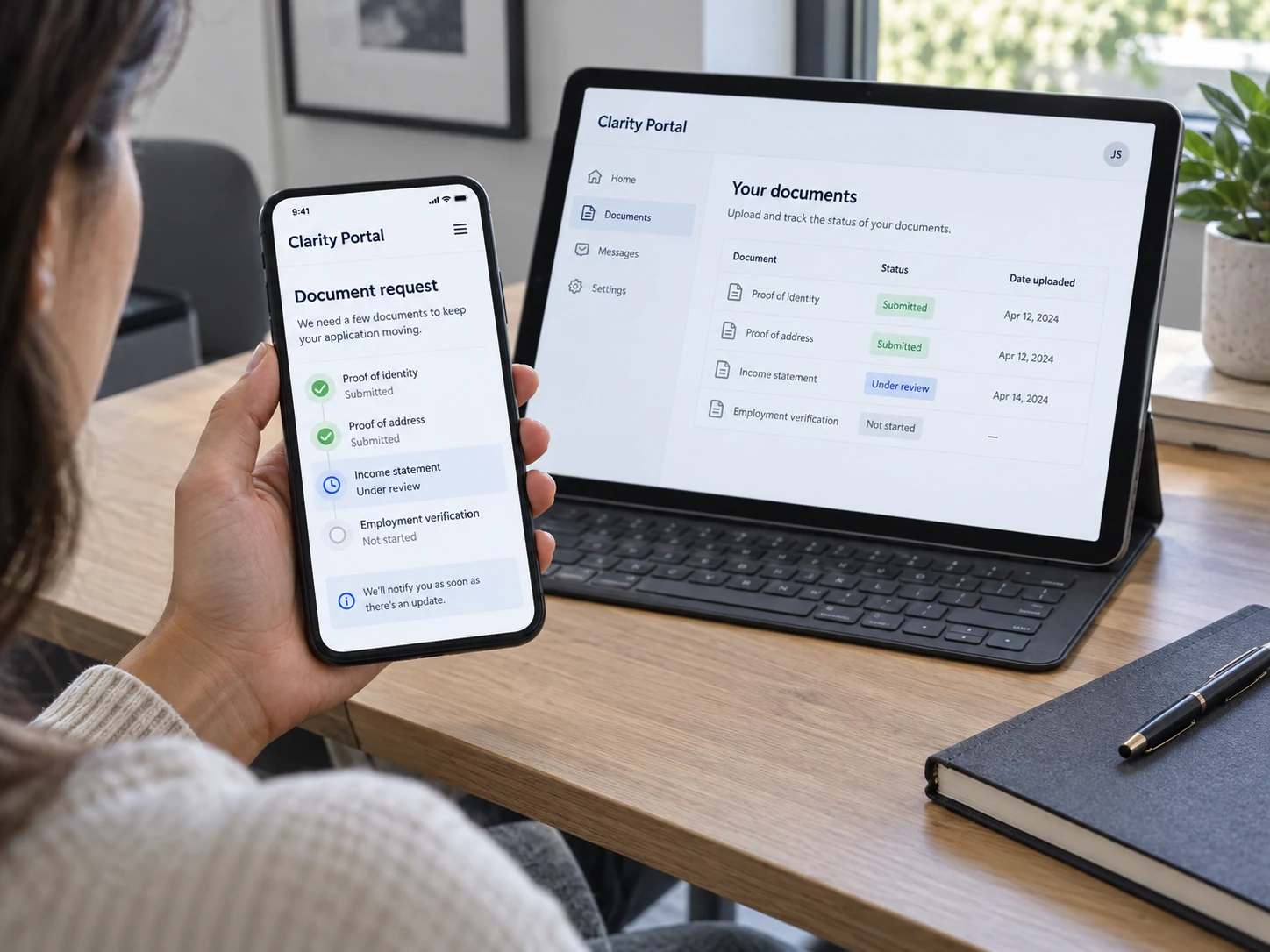 Seated customer at a desk uses a phone supported in left palm to review fictional 'Clarity Portal', clear document request and 'Under review' status, landscape tablet nearby shows matching document list
