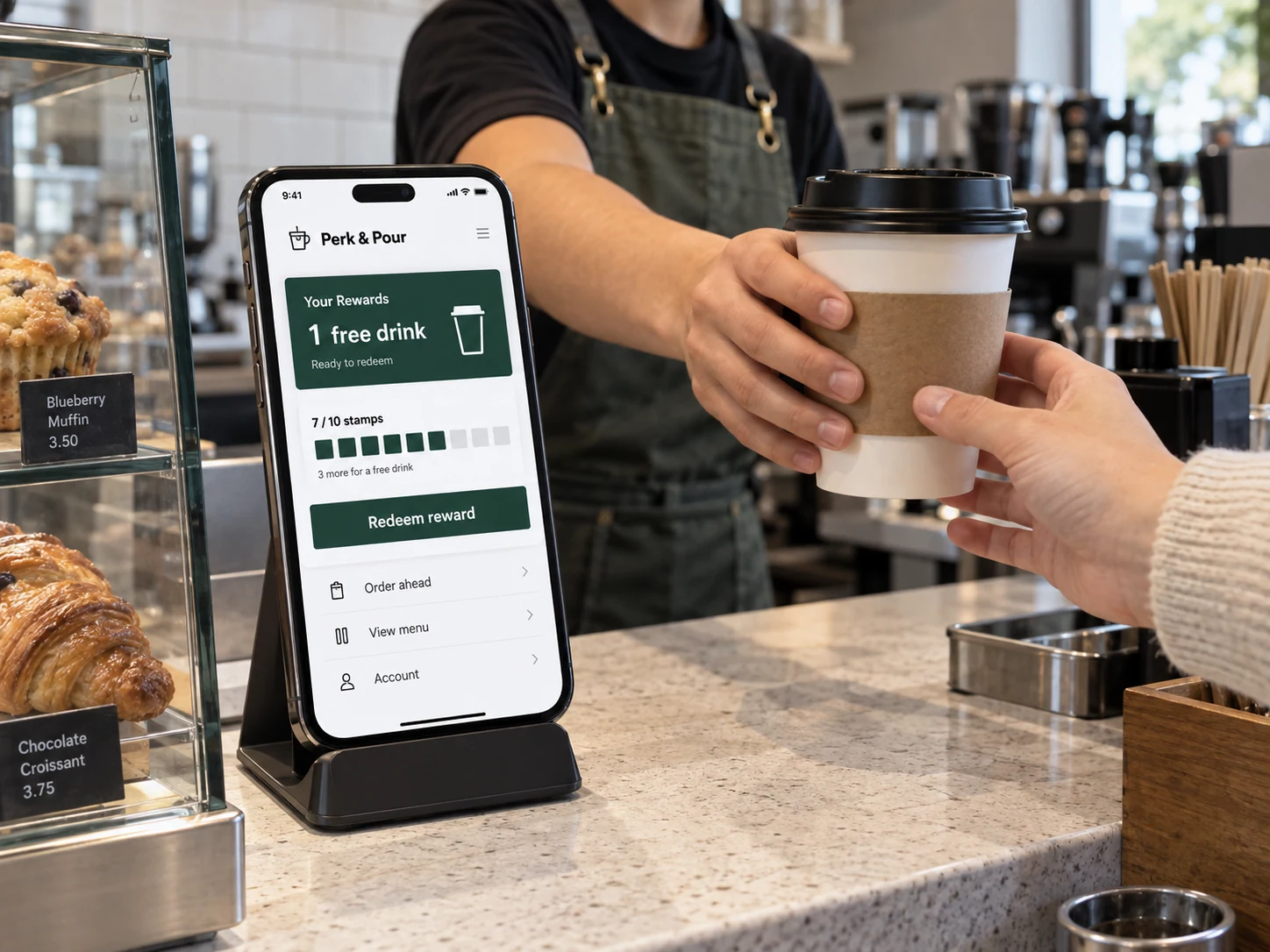 Smartphone on café counter stand displays fictional 'Perk & Pour' loyalty app with clear reward card and 'Redeem reward', barista hands pass a coffee cup beside the screen